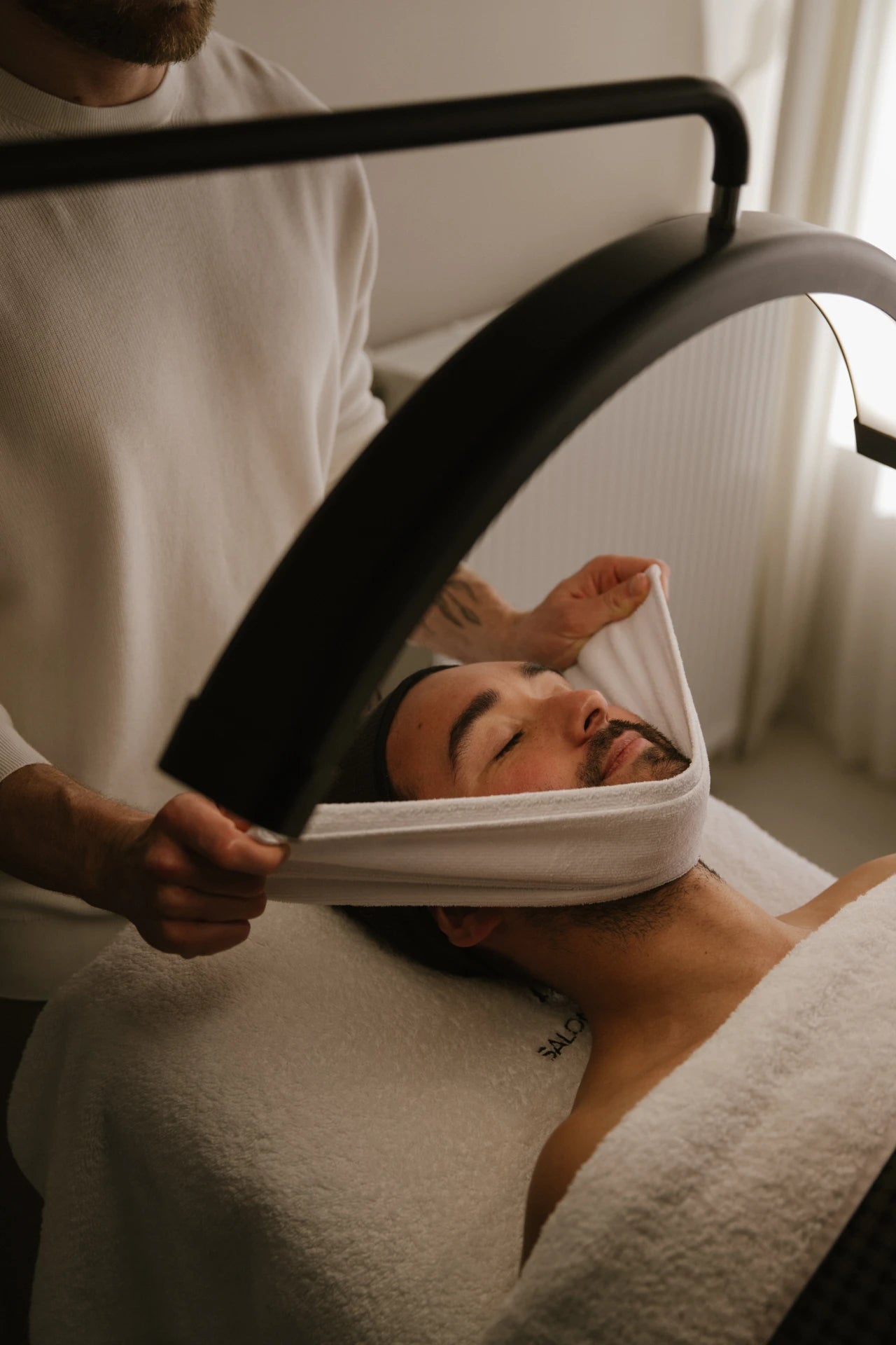 A warm towel being carefully wrapped around a client’s face during a relaxing skincare treatment at Salon d’Homme.